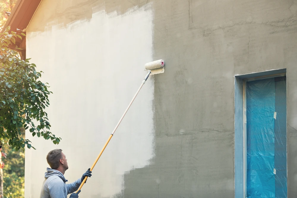 Un homme peint un mur extérieur beige avec un rouleau à manche télescopique