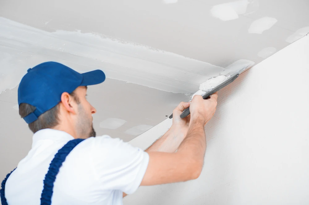 Artisan en tenue de travail blanche et casquette bleue applique de l'enduit au couteau sur un mur intérieur blanc près du plafond