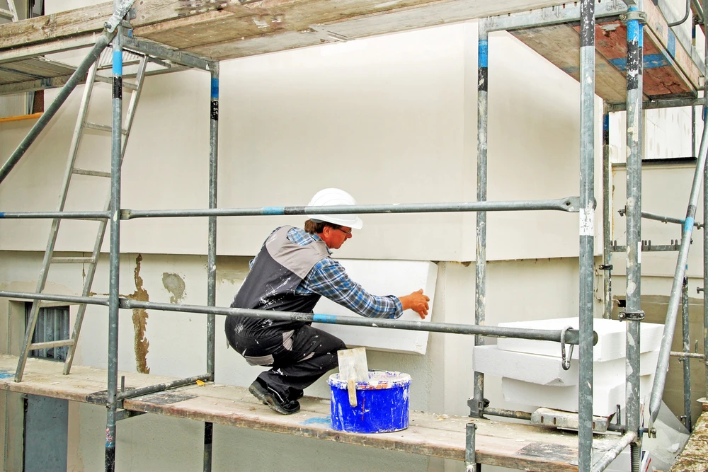 Ouvrier du bâtiment en vêtements de travail et casque blanc appliquant un enduit ou peinture blanche sur façade extérieure depuis échafaudage métallique