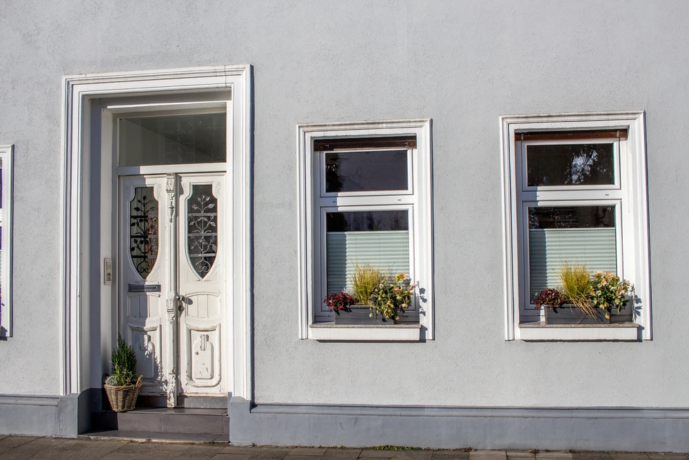 Façade extérieure d'habitation avec porte d'entrée blanche ornée de vitraux décoratifs et deux fenêtres à volets roulants partiellement fermés
