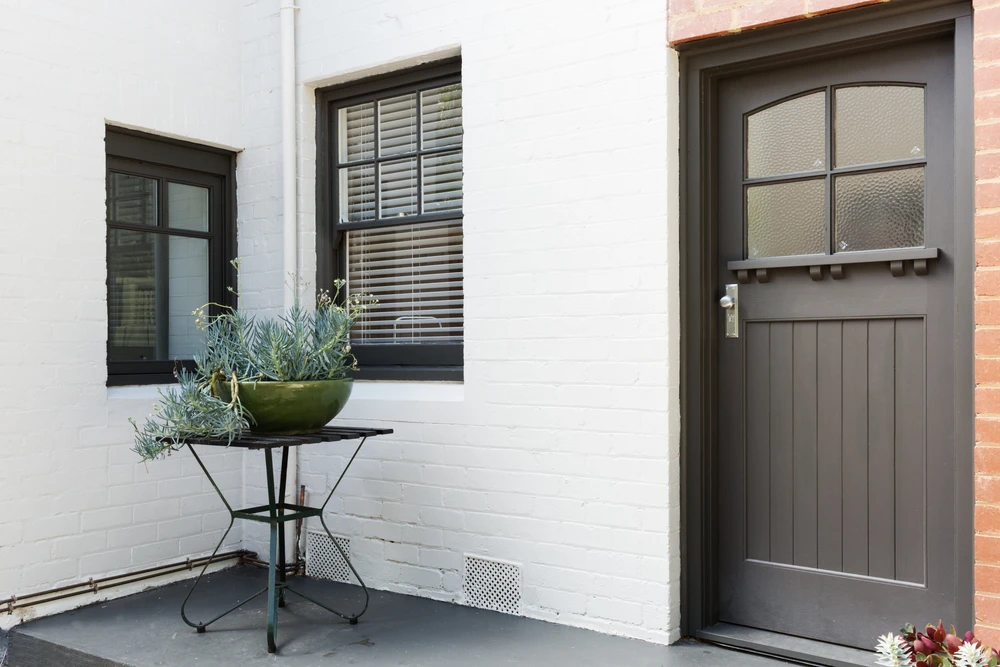 Entrée extérieure d'une maison avec mur blanc en briques peintes, porte d'entrée grise à panneaux avec vitrage texturé et poignée chromée