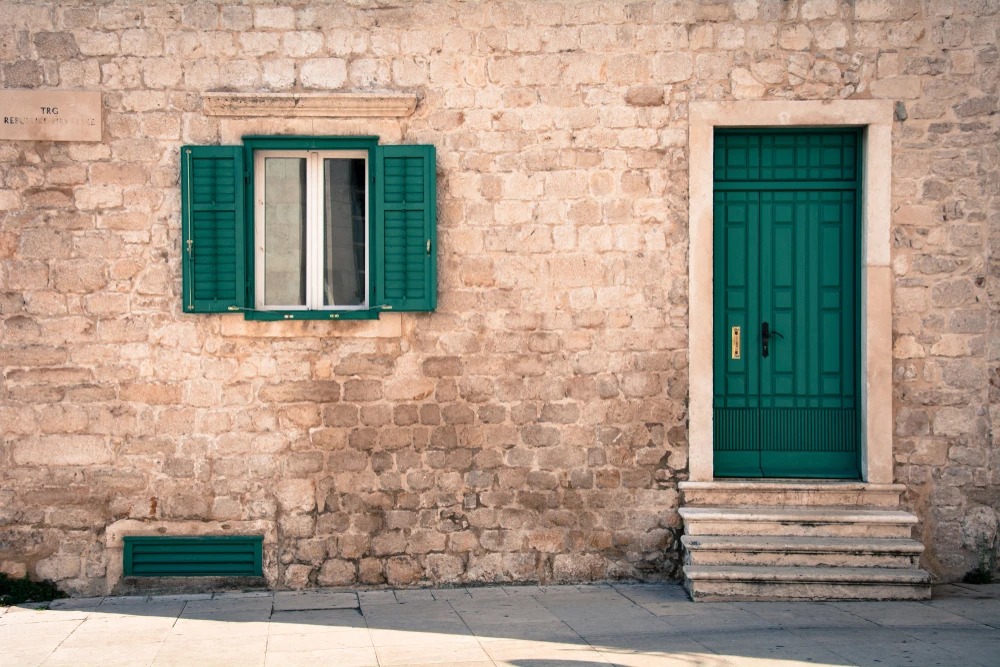 Façade extérieure d'une maison traditionnelle en pierre calcaire avec porte d'entrée verte à panneaux et fenêtre assortie aux volets ouverts