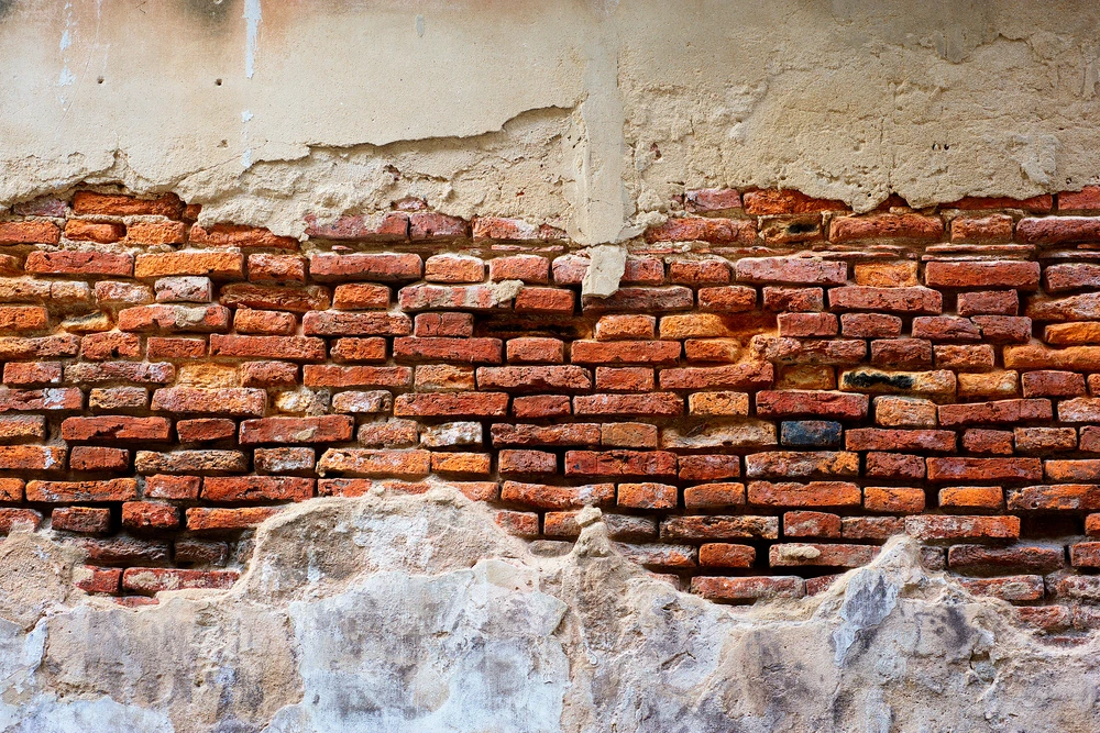 Mur extérieur ancien en détérioration montrant une maçonnerie de briques rouges apparentes sous un enduit de façade écaillé et fissuré