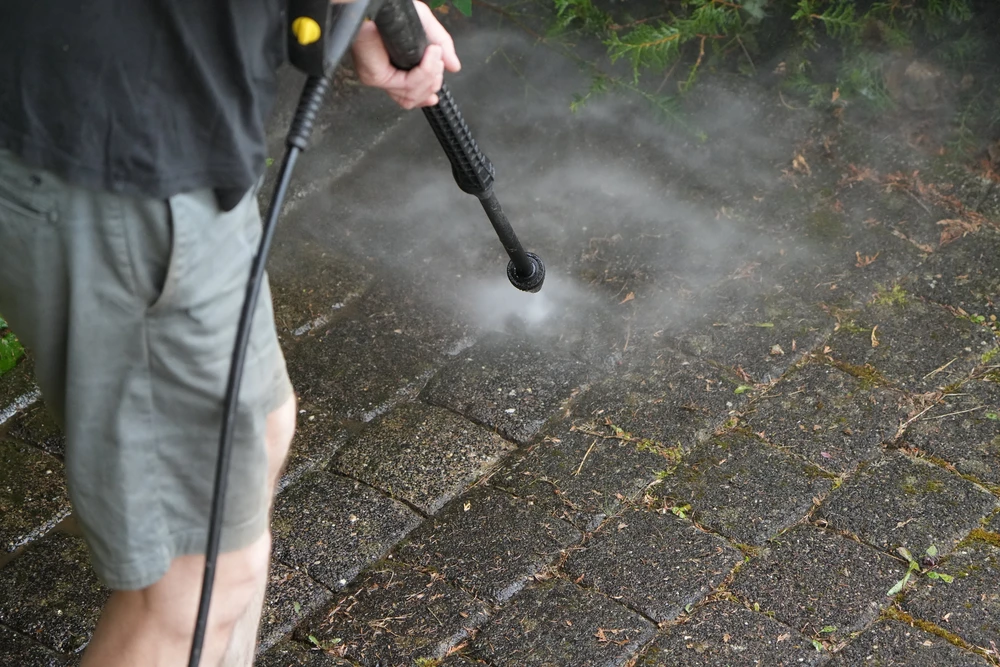 Homme utilisant un nettoyeur haute pression sur pavés extérieurs, projection de vapeur d'eau visible, élimination mousses et saletés accumulées entre joints
