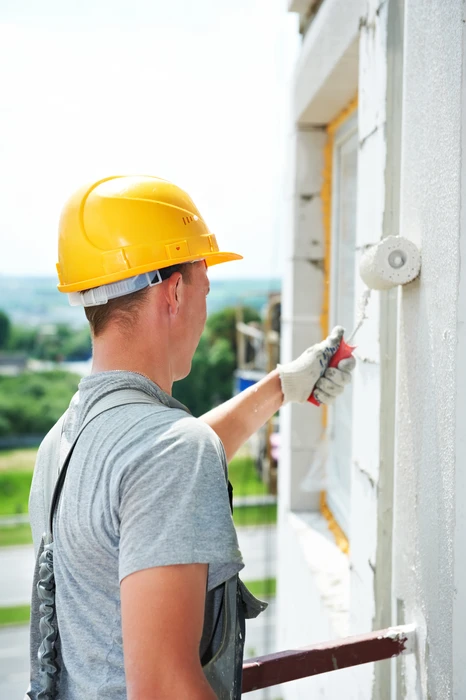 Ouvrier du bâtiment en casque jaune applique un enduit ou peinture blanche sur façade extérieure avec rouleau