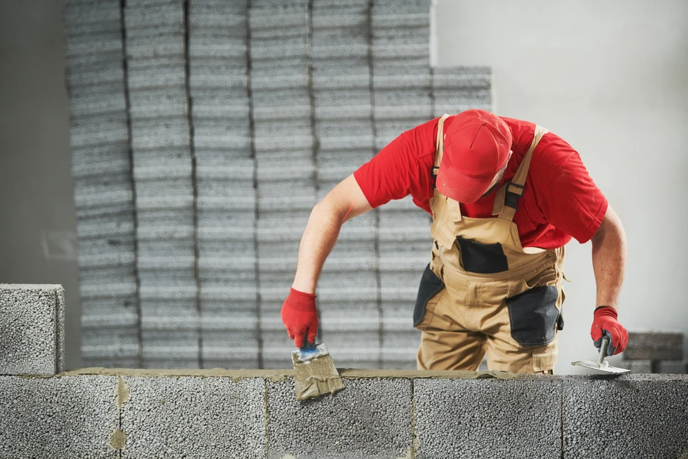 Maçon en tenue de travail rouge et salopette beige applique du mortier sur mur de parpaings à l'aide d'une truelle