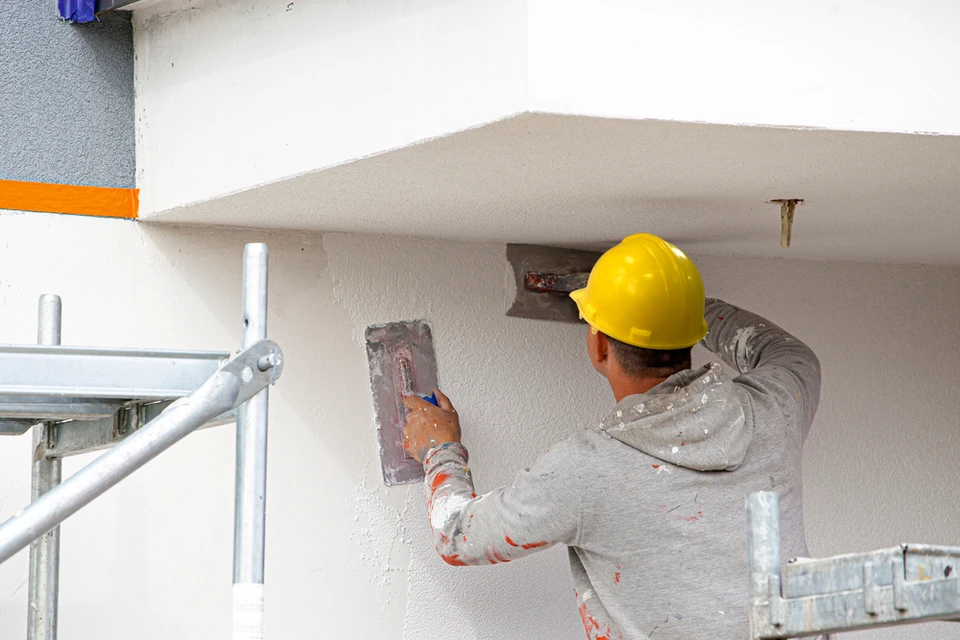 Ouvrier en bâtiment appliquant un enduit sur façade extérieure, portant casque de sécurité jaune et vêtements de travail